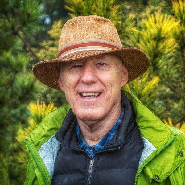 Smiling older man, John J. Albers, in a brown hat and green jacket outdoors with greenery behind.