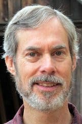 Alex Wilson, is a man with gray hair and a beard. He stands outdoors, smiling and wearing a maroon shirt.