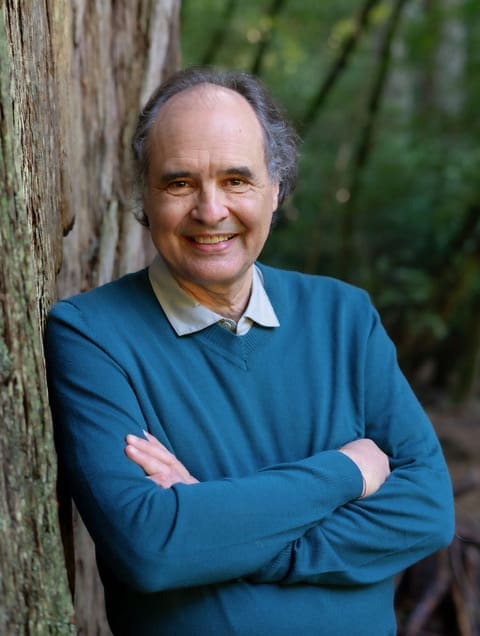 Smiling Andrés Edwards in blue sweater stands with arms crossed by a tree in a forest.