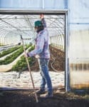 The Winter Market Gardener stands with a rake before a lush, green-filled greenhouse.