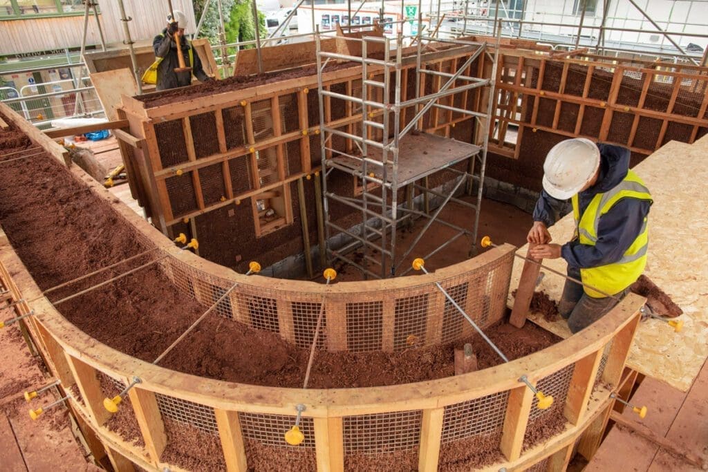 A worker in a white hard hat kneels on a section of plywood as he sets a wall post of a cob structure above the ground.
