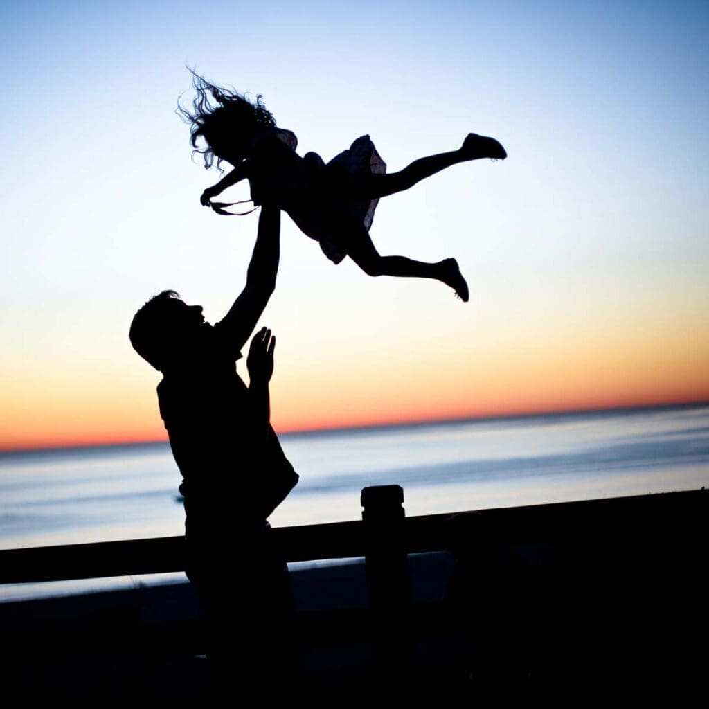 The silhouette of a man holding a young child over his head next to the ocean as the sun sets on the horizon.