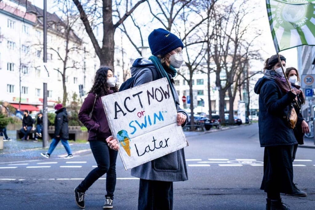 Masked activists demonstrate on a city street; one holds an Act Now or Swim Later sign.
