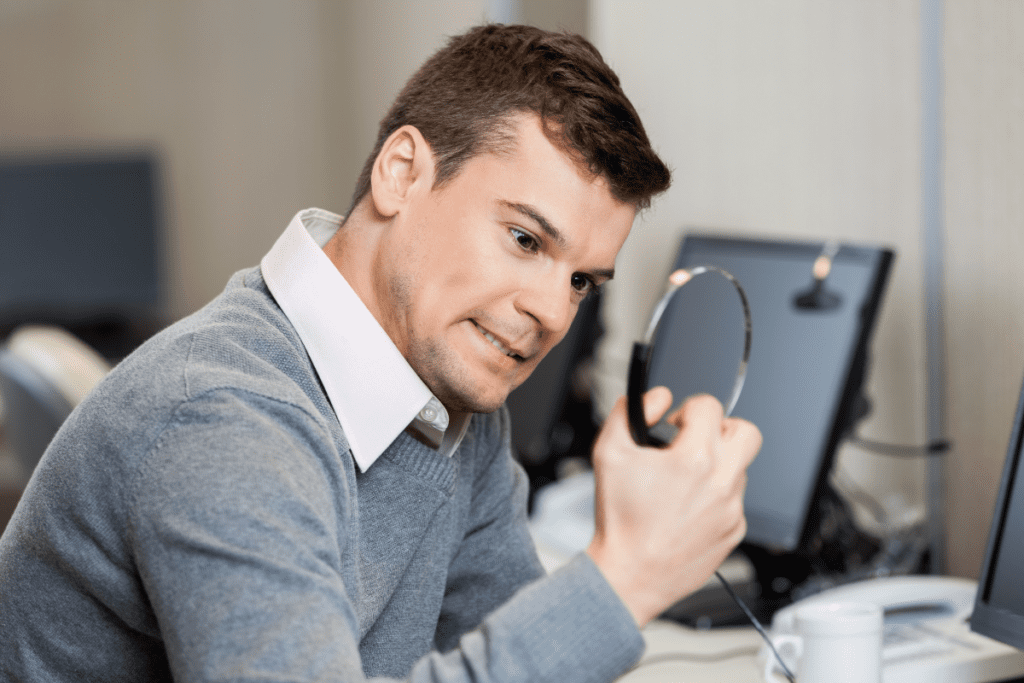 A young man in an office holds a computer headset in his fist as he stares angrily down at it with a scowl on his face.