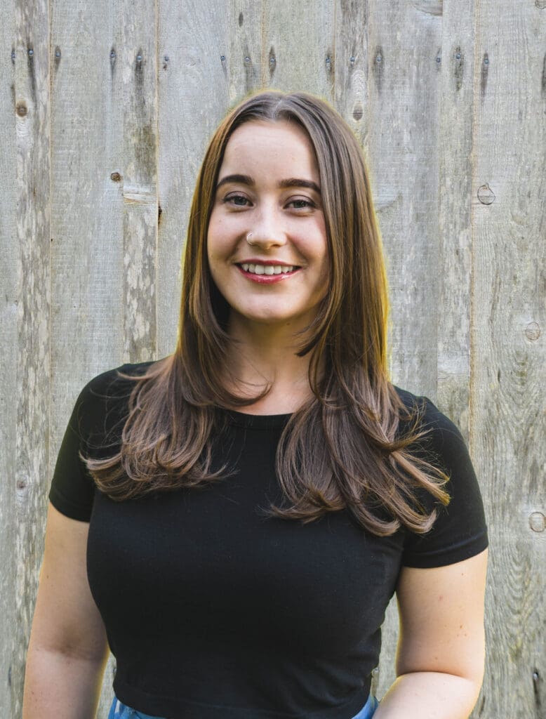 Molly Harrison, with long brown hair and a black top, smiles in front of a wooden fence.