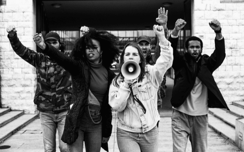 A black-and-white image of a group of young protesters raising their fists and yelling (one into a microphone)