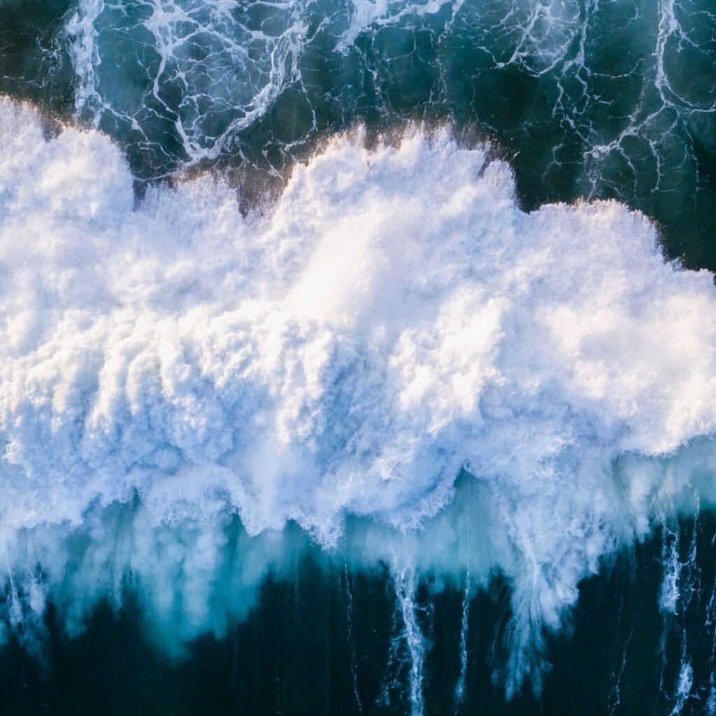 Aerial view of ocean waves crashing against each other, creating a frothy white foam. The deep blue water contrasts with the vibrant foamy crests, showcasing the dynamic and powerful movement of the sea.
