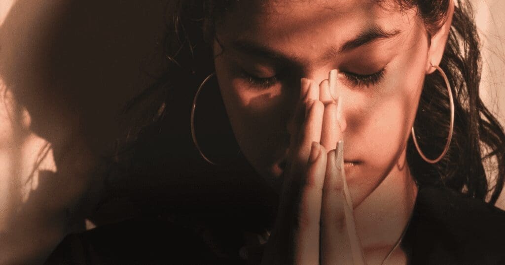 A young woman stands in shadow with her hands held to her face