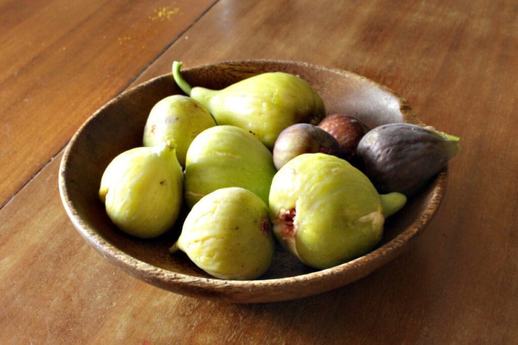 A Figs-in-bowl, brimming with ripe figs in a spectrum of colors—a mix predominantly of yellowish-green and some purplish hues—rests on a wooden table. Even though figs thrive in warmer climates, you can cultivate them in colder areas by shielding them from frost. The textures of both the figs and the bowl are distinctly visible.