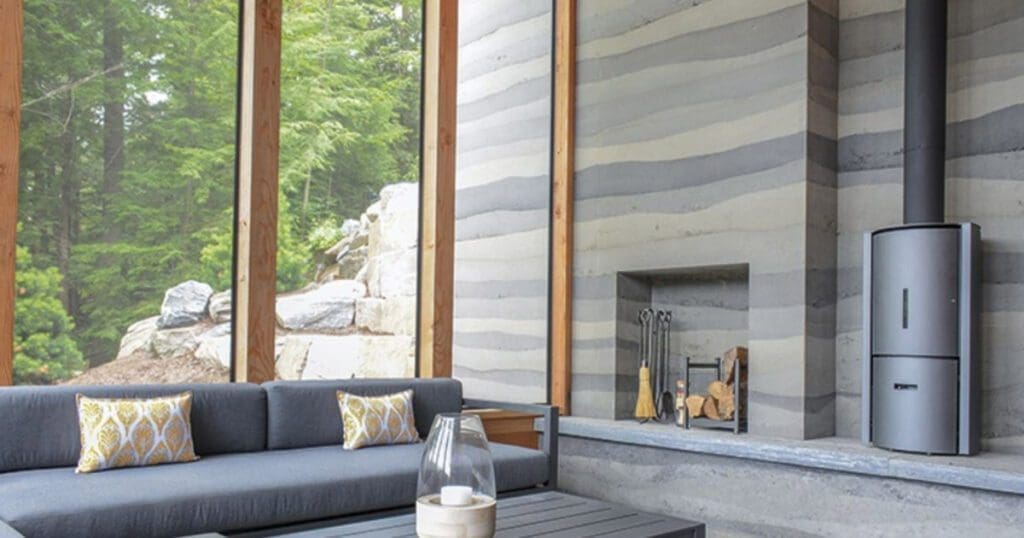 A modern living room with large floor-to-ceiling windows showcasing a wooded outdoor view features a gray sectional sofa adorned with yellow-patterned throw pillows. The room is complemented by a coffee table displaying decorative items and a sleek metal wood-burning stove positioned beside an area stacked with firewood, all of which emphasize the concept highlighted in the Social_Excerpt_building-with-materials-found-onsite.