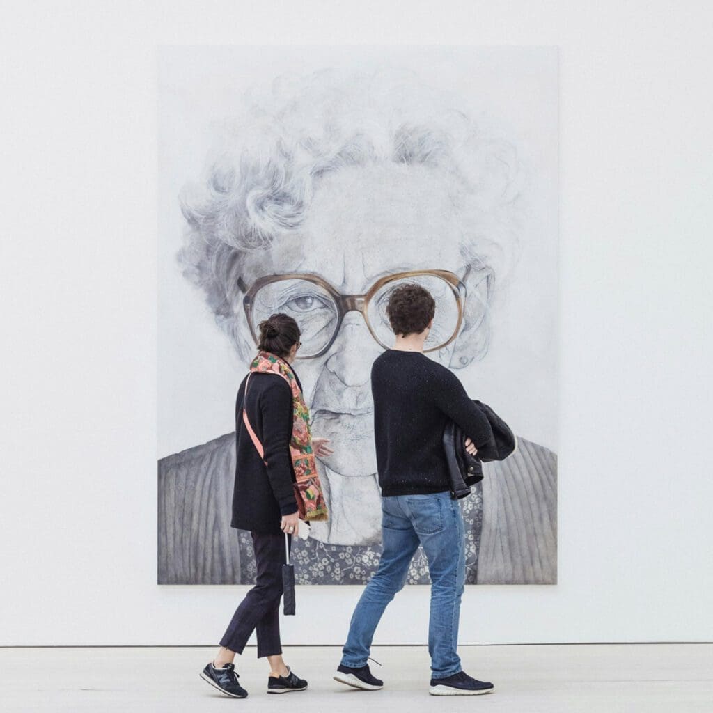 Two individuals are walking past "ryan-stefan-5K98ScREEUY-unsplash," a large, gray-toned portrait of an elderly person with curly hair and large glasses. The portrait is displayed on a white wall. One person is wearing a black top and colorful scarf, while the other in a black sweater, quietly discussing climate change.