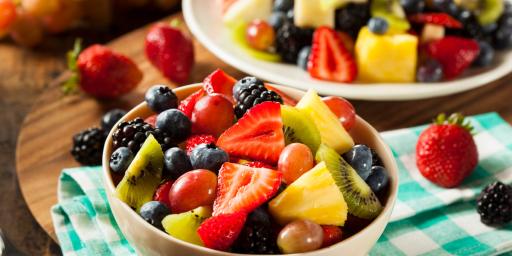 A bowl of better, organic fruit salad with strawberries, kiwi, blueberries, and pineapple sits on a checkered cloth.