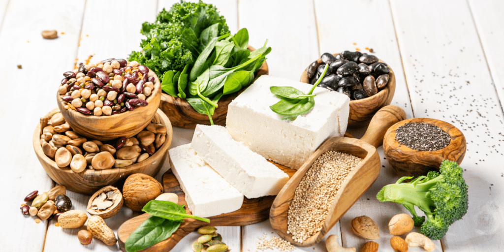 A variety of organic plant-based foods, including tofu, beans, seeds, nuts, and leafy greens on a table.