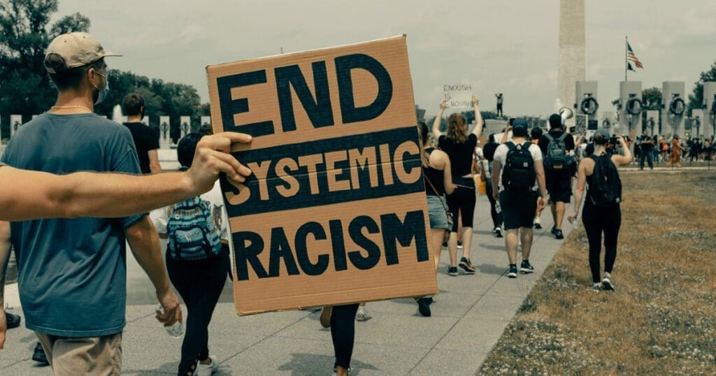 Protesters march holding a sign that reads "End Systemic Racism" near a monument, sparking vital communication on race.