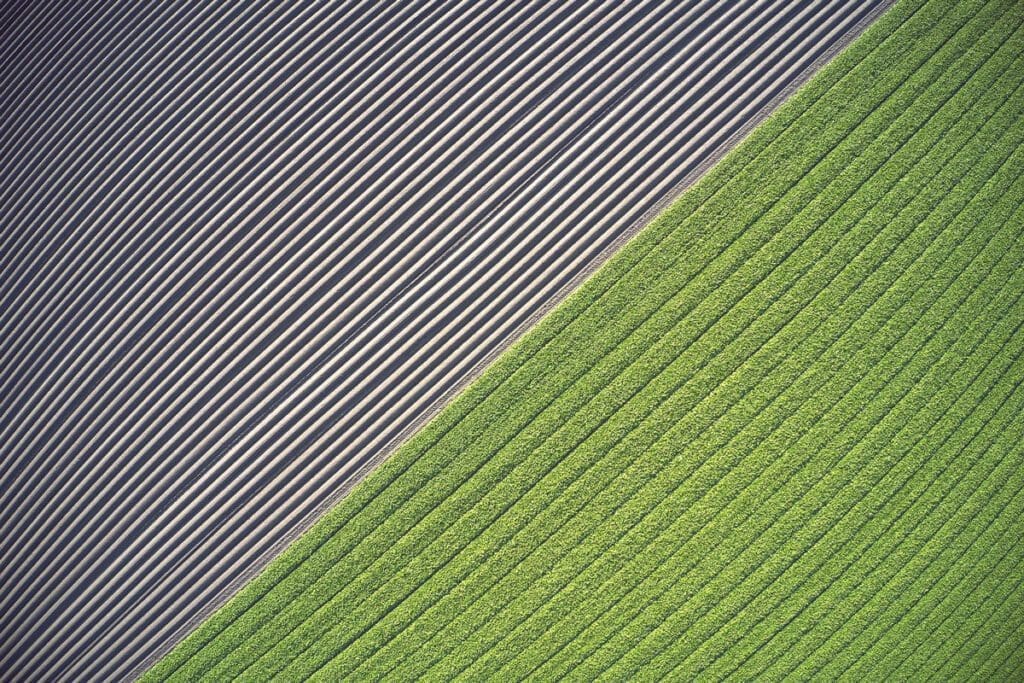 An aerial view reveals a healthy landscape with a stark contrast between plowed fields and vibrant green crops, divided diagonally.