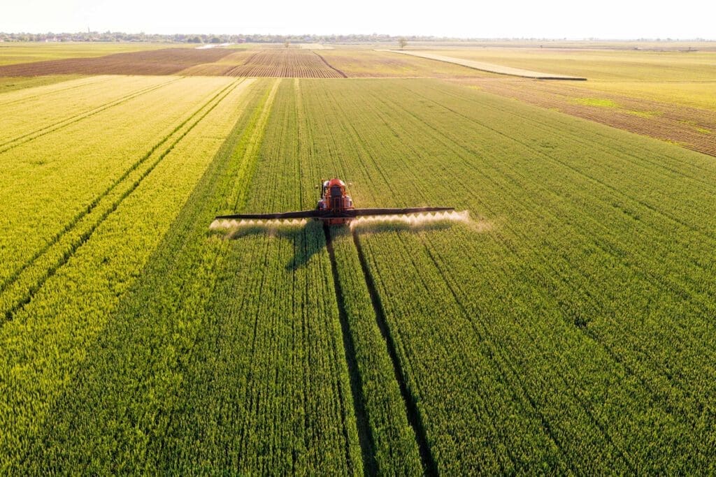 Aerial view of a tractor spraying crops in a vast green field under a clear sky, contributing to a healthy food system that nurtures our planet.