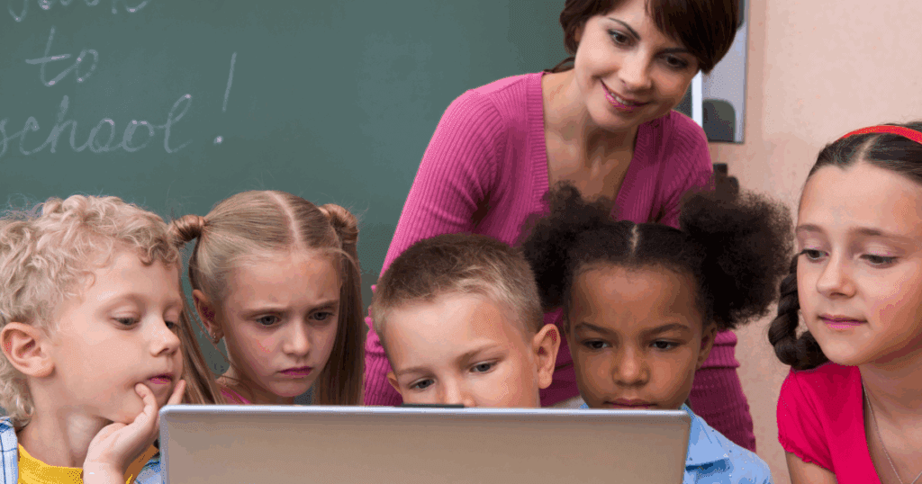 Teacher and five young children looking at a laptop together in a classroom.