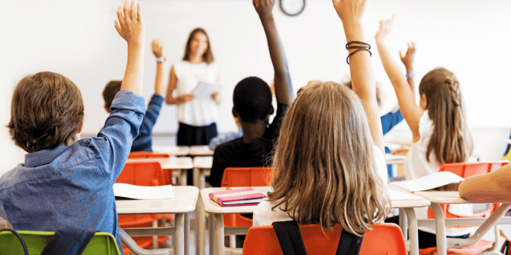 Students sitting at desks raise their hands in a classroom while a teacher stands in front.