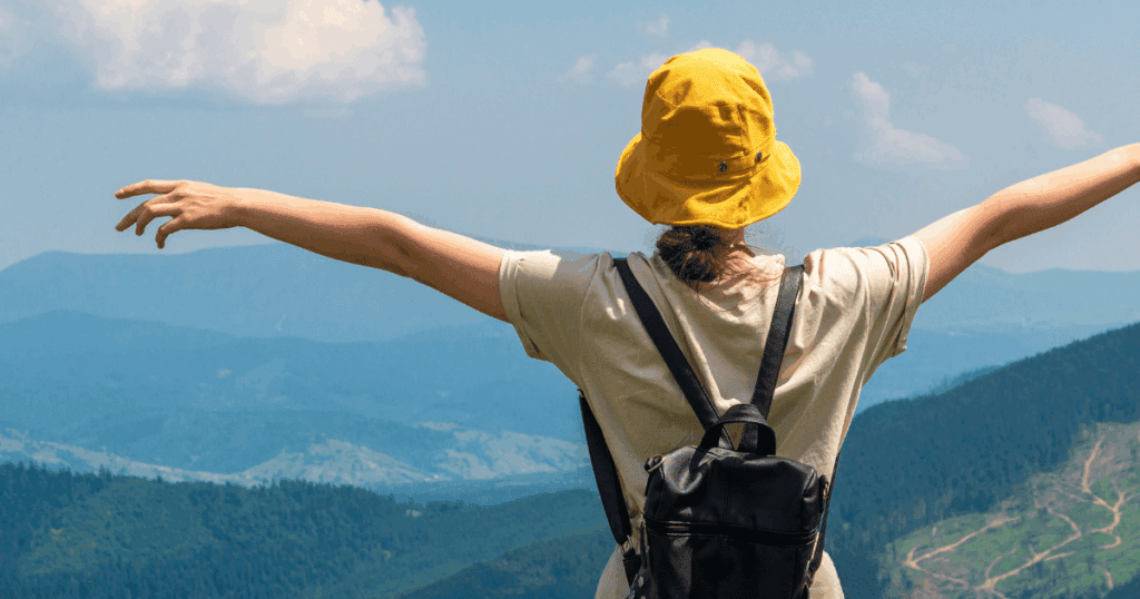 Person with a yellow hat and backpack enjoys the summer outdoors atop a mountain, arms outstretched.