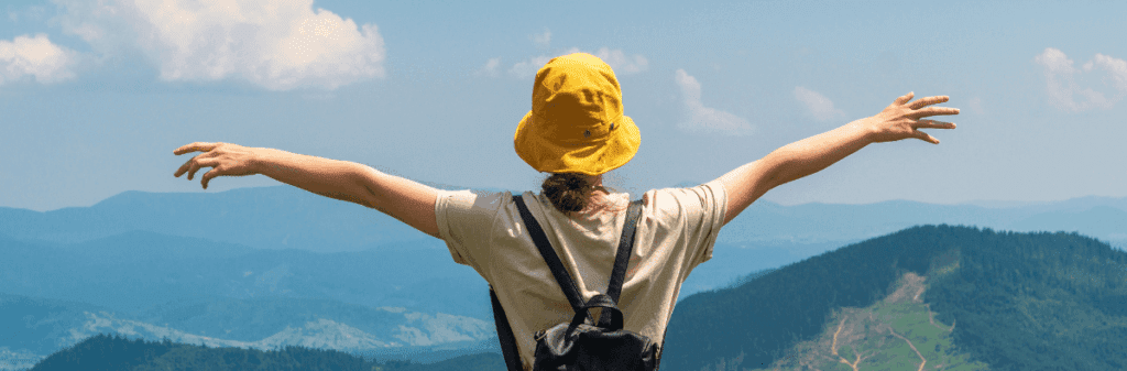 Person in yellow hat with arms outstretched enjoying the summer outdoors, overlooking mountains.