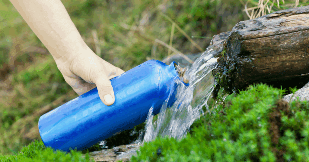 Hand filling a blue water bottle from a natural stream in the outdoors during summer.