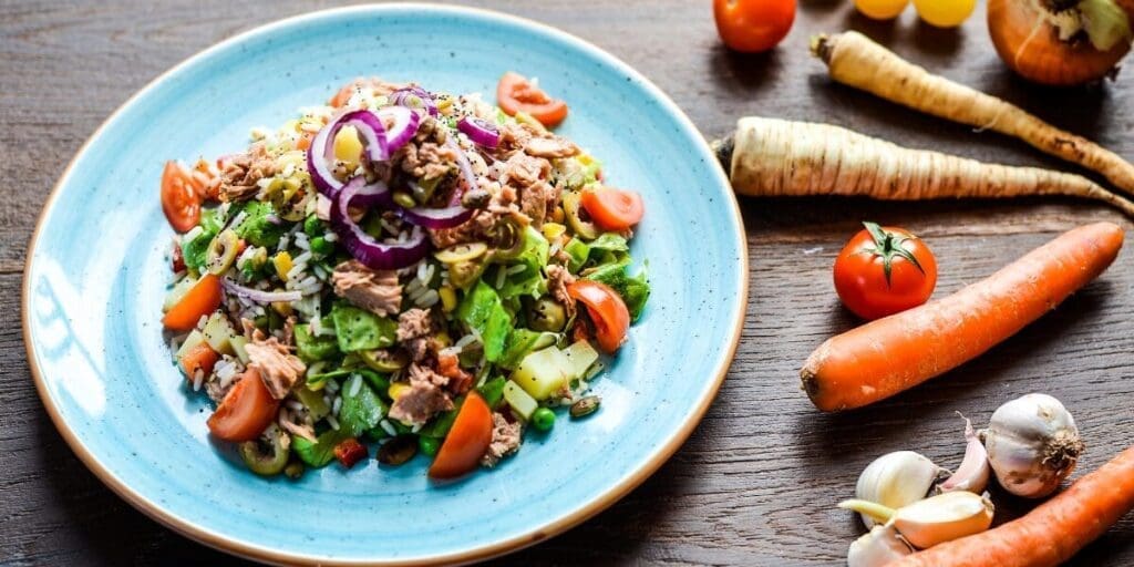 Colorful salad with tuna, onions, veggies, and nuts on a blue plate, surrounded by fresh produce.