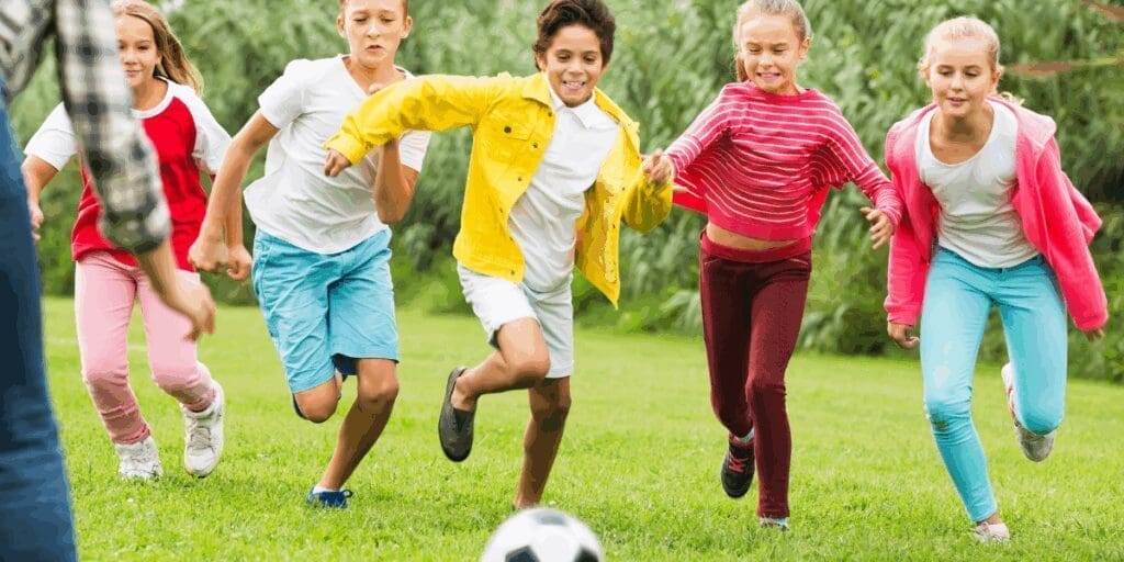 Five kids chase a soccer ball on a grassy field, smiling and running with summer joy.