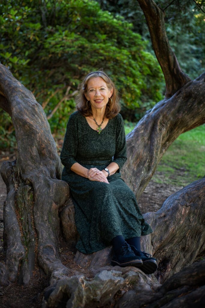 Marlene Schiwy in a green dress sits on a large tree branch in a lush, green outdoor setting.