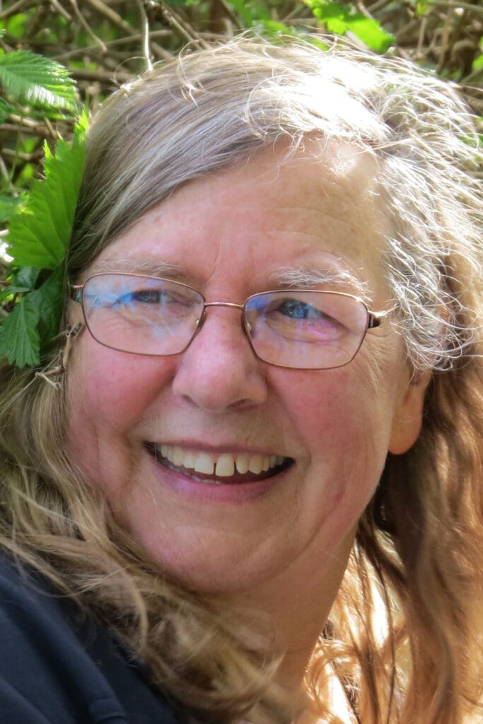 Smiling older woman, Nancy Turner, with glasses and long hair, outdoors with green leaves in her hair.
