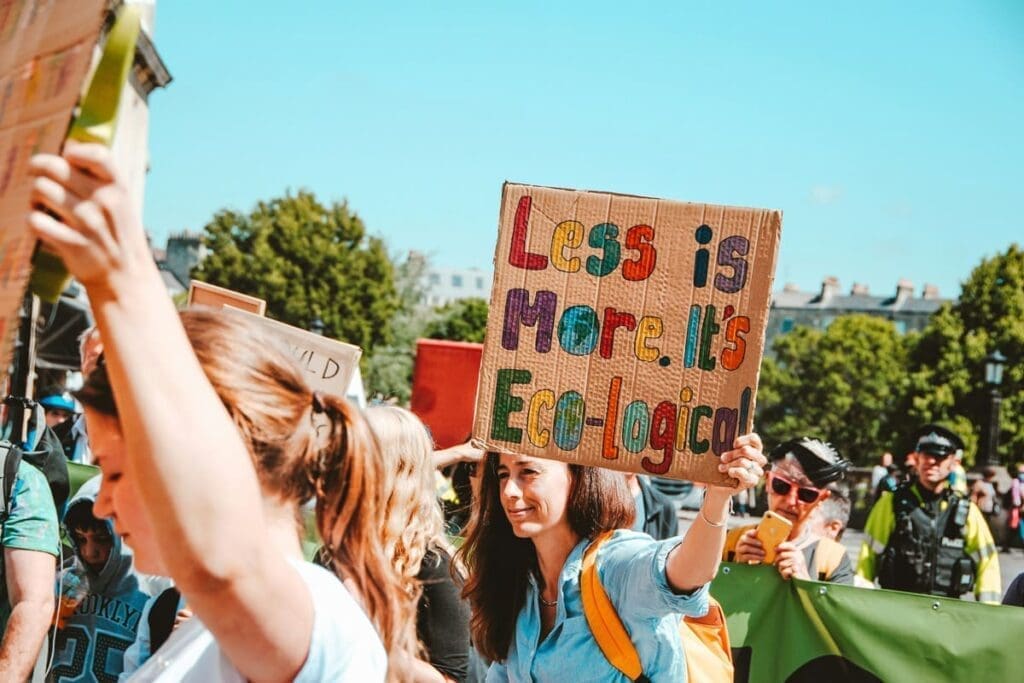 Woman holds sign reading "Less is More. It's Eco-logical! Aim for 2.5 tonnes carbon math.