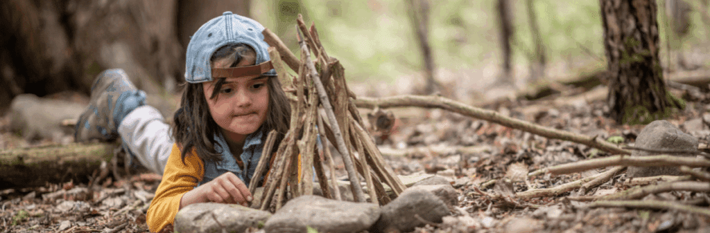 A timeless scene: a child in a cap builds a small teepee with sticks in the forest.