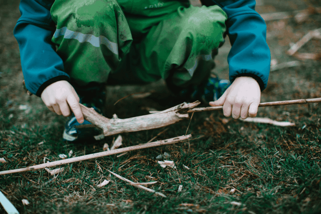 Child in green pants breaking sticks on grass, enjoying outdoor play.