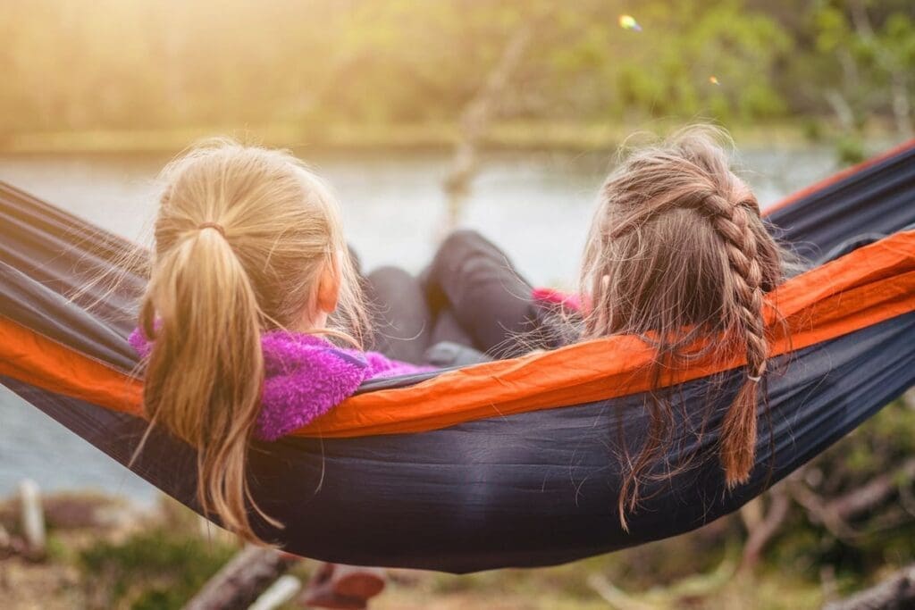 Two girls relax in a hammock by the water, talking about climate change on a sunny day.