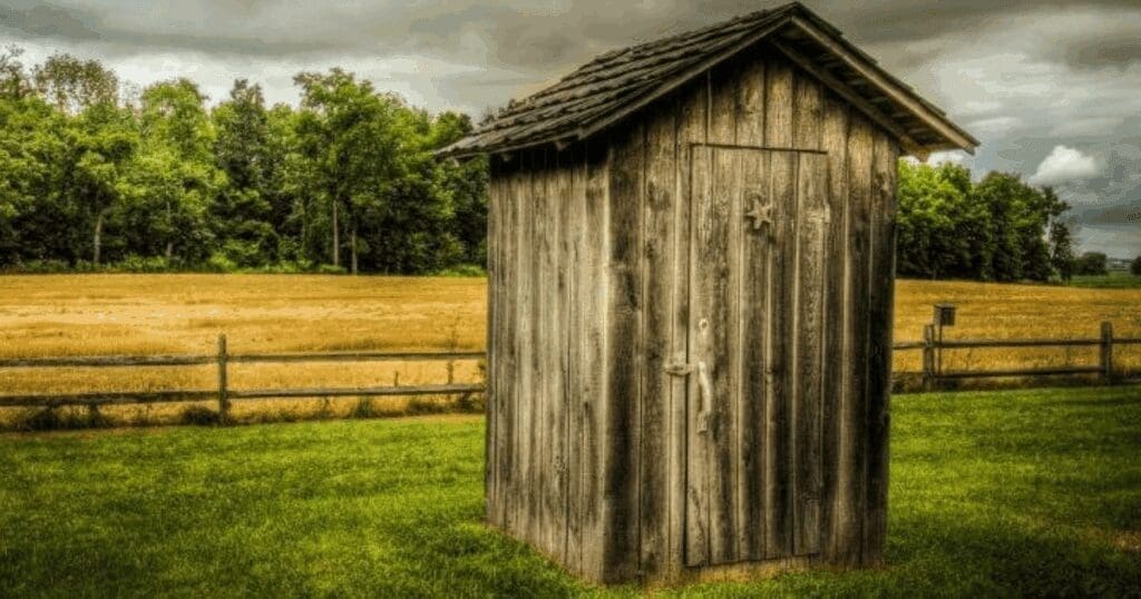 A weathered wooden outhouse stands on grass, reflecting Gord Baird’s composting toilets ethos.