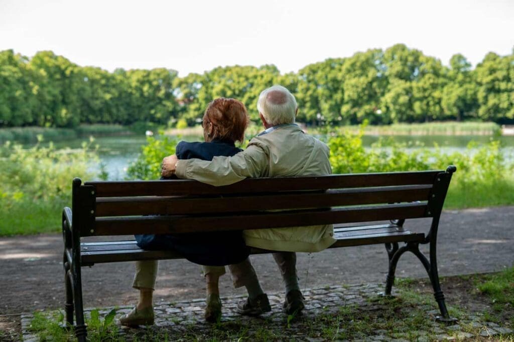 An elderly couple sits arm in arm by the lake, enjoying the peaceful community commons.