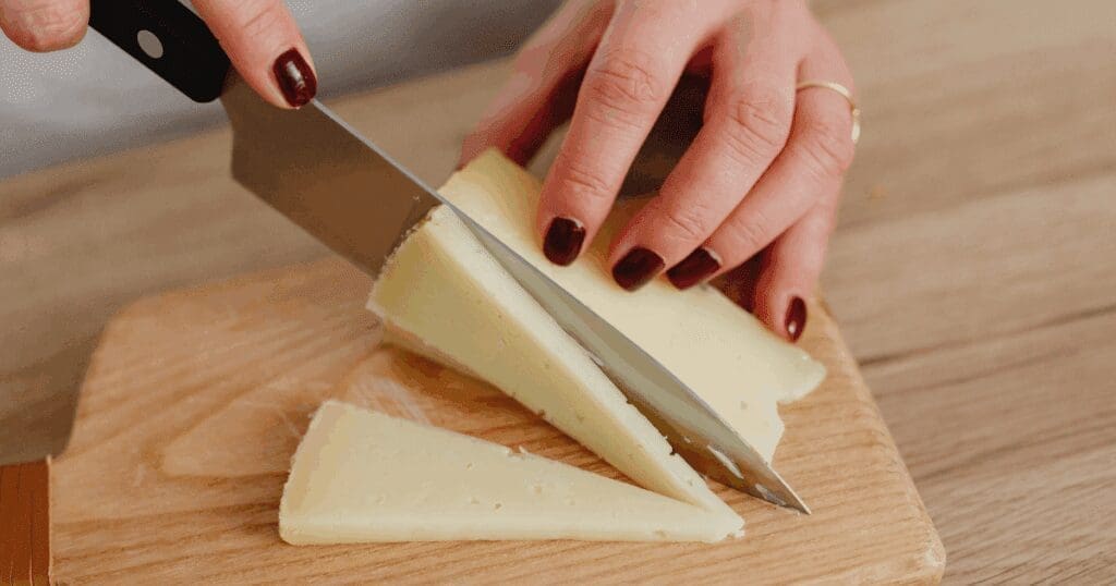 A person slicing homemade cheese into triangles on a wooden cutting board with a large knife.