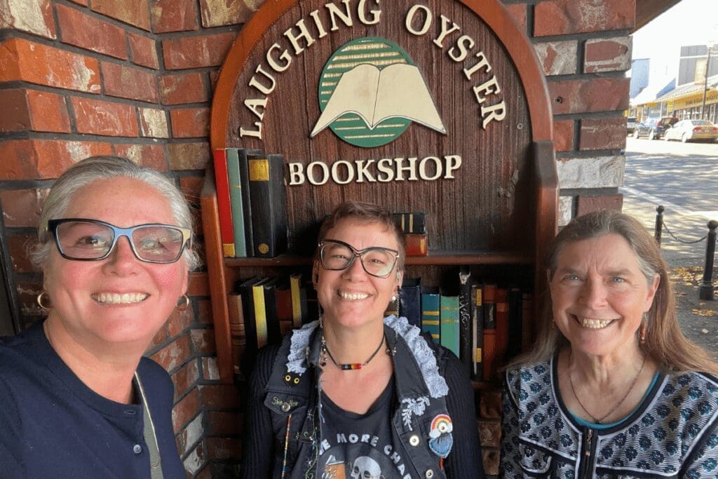 Three smiling women stand outside Courtenay’s independent Laughing Oyster Bookshop on a sunny day.