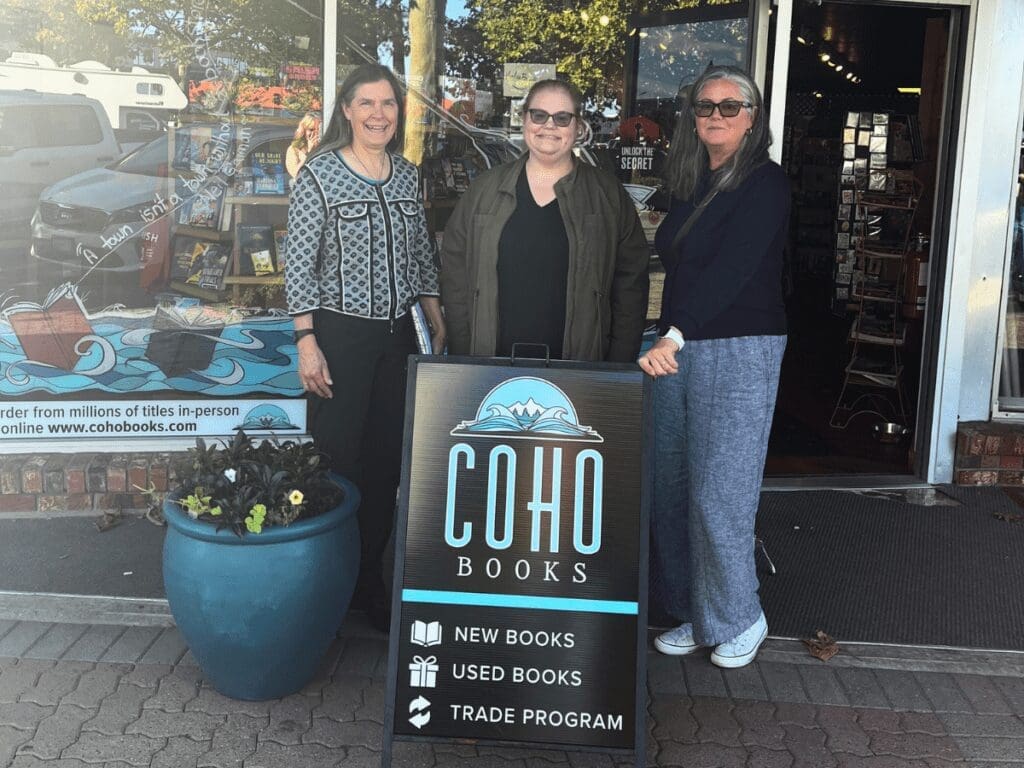 Three women stand downtown by Coho Books, next to a sign for Curious Readers programs.