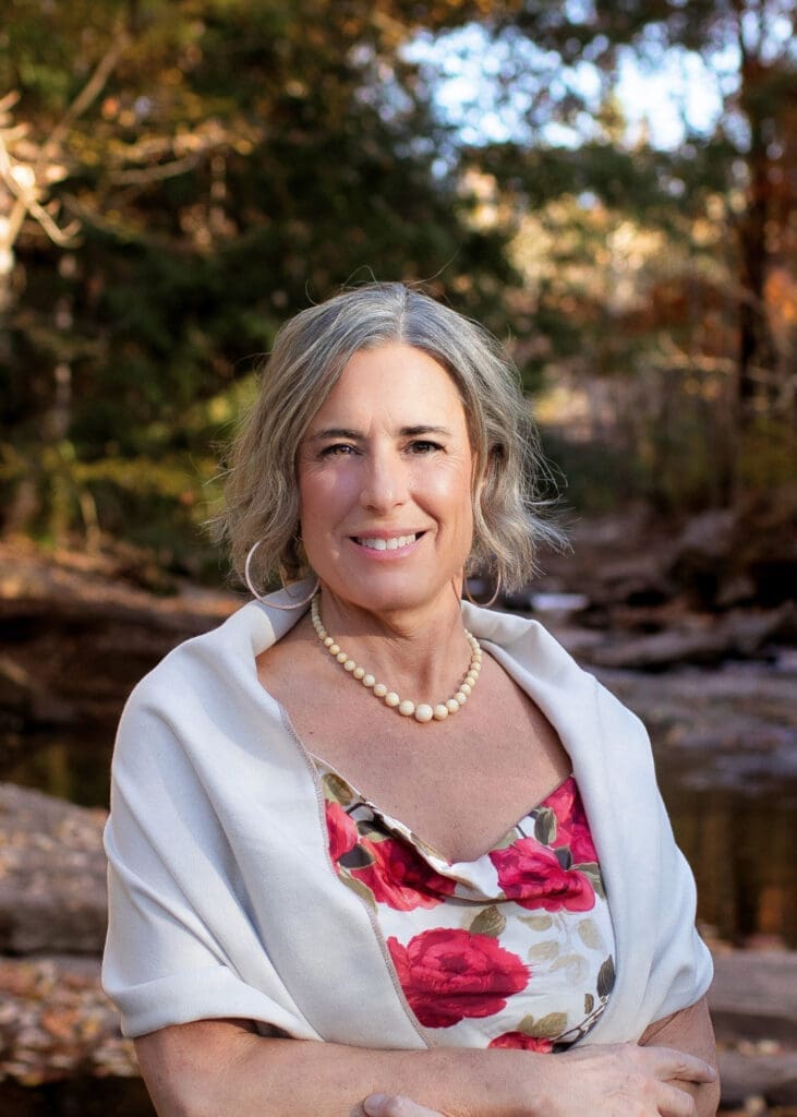 Smiling woman in floral dress and pearl necklace standing outdoors near a creek and trees.