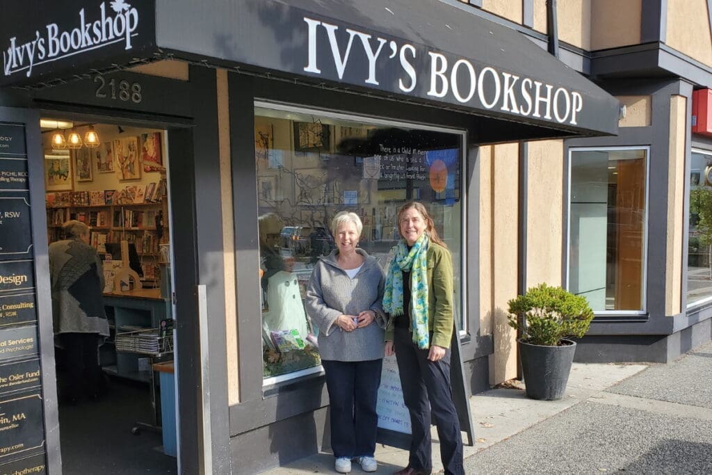 Two women stand outside Ivy’s Bookshop downtown, smiling at the camera on a sunny day.