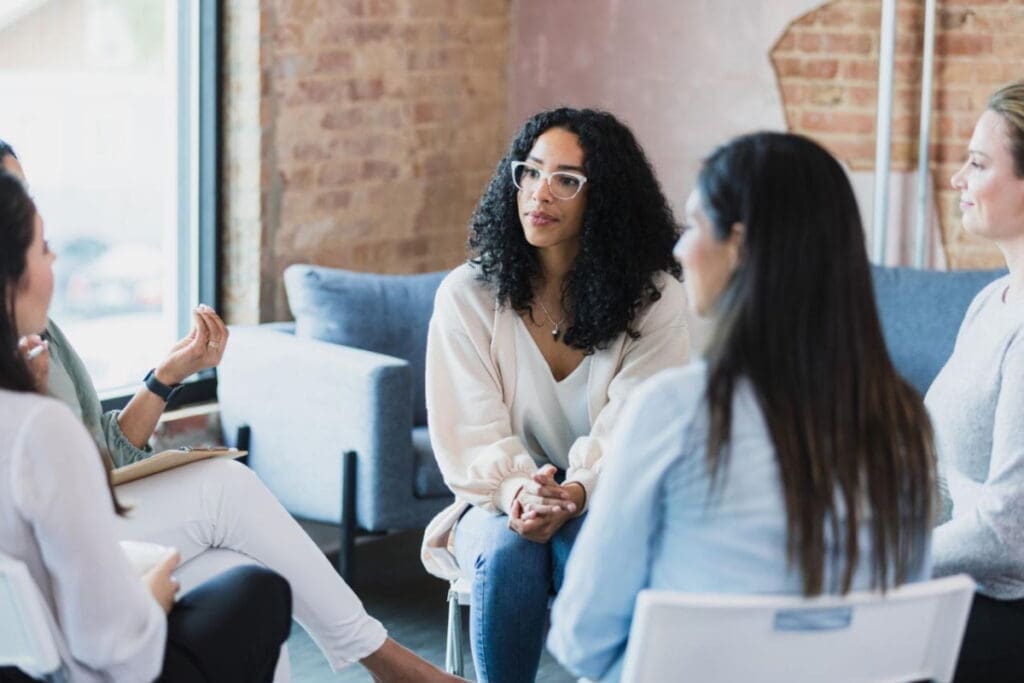 Four women sit in a circle as a workshop facilitator holds space for group discussion indoors.