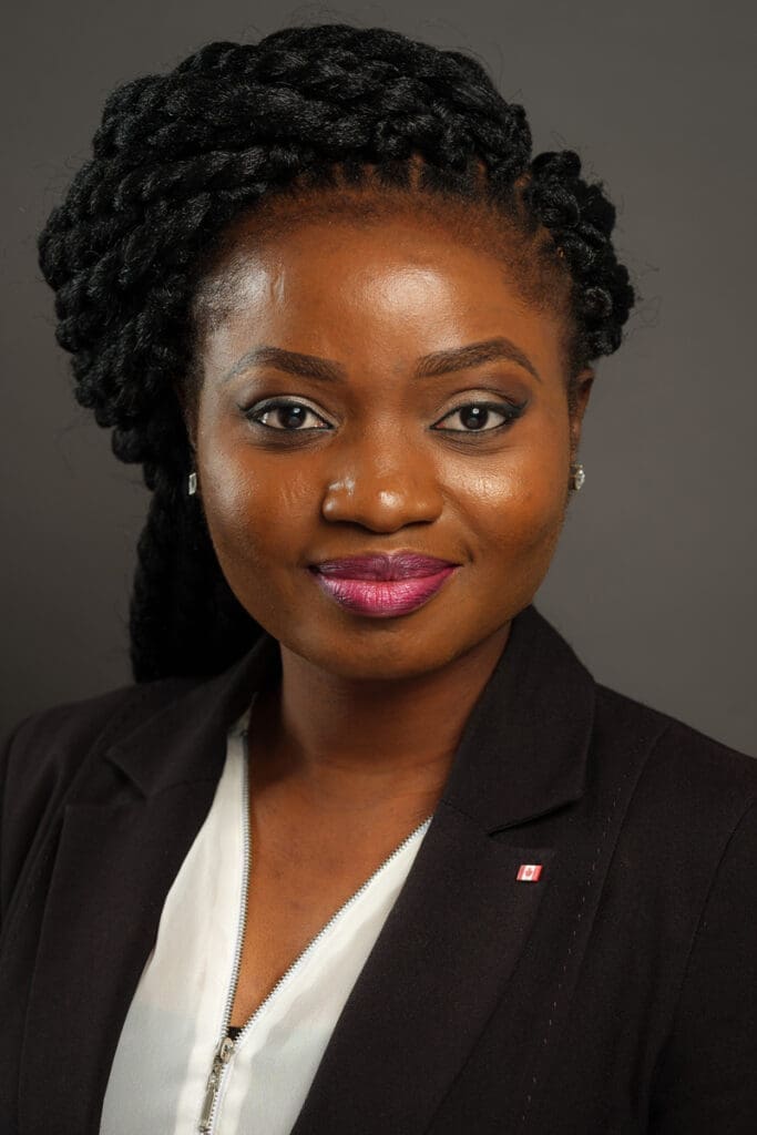 Ruth Mojeed Ramirez, with braided hair, wears a black blazer and white blouse, smiling confidently.