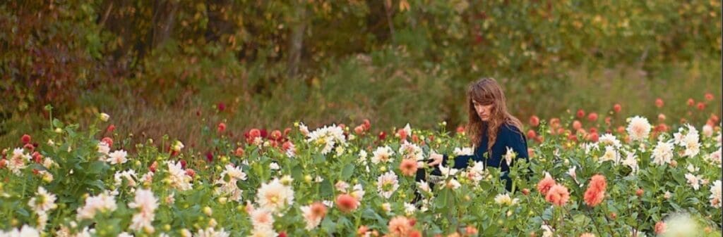 A person stands among blooming flowers, embracing cut flower season in a field with trees behind.