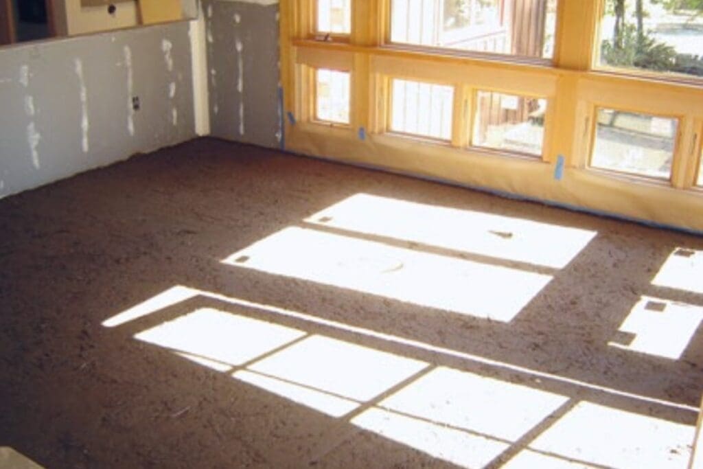 Room with unfinished walls and earthen floor, sunlight streaming through large windows.