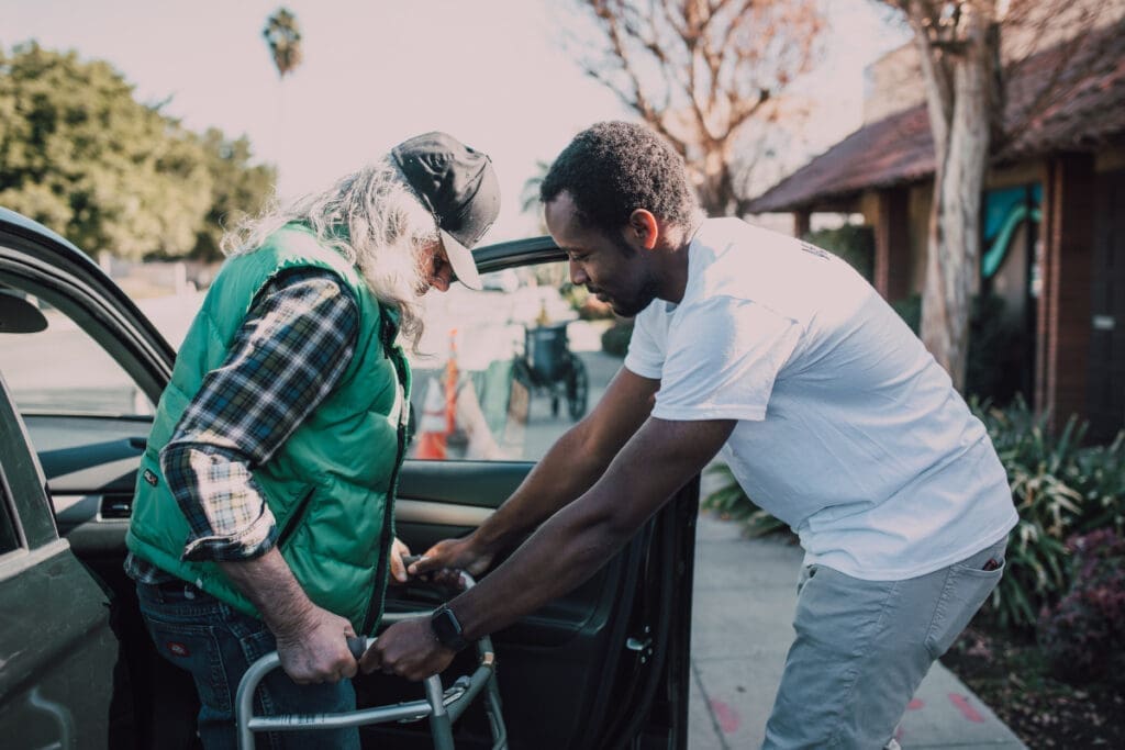 Man helps older person with walker out of car, embodying transformation on a sunny day outdoors.