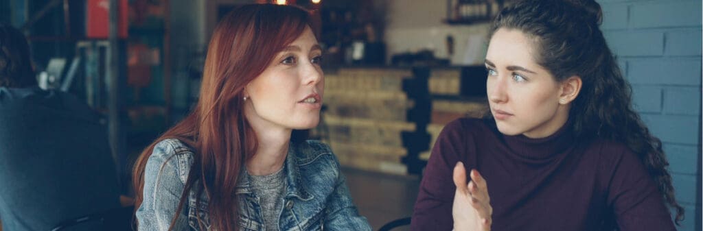 Two women sit at a cafe table, engaged in a serious conversation about conflict resolution.