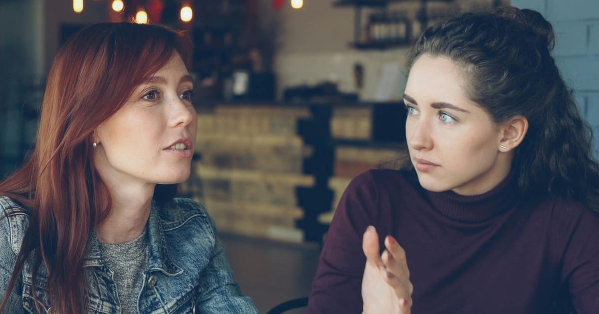 Two women indoors, sharing a serious conversation about conflict resolution at a table.